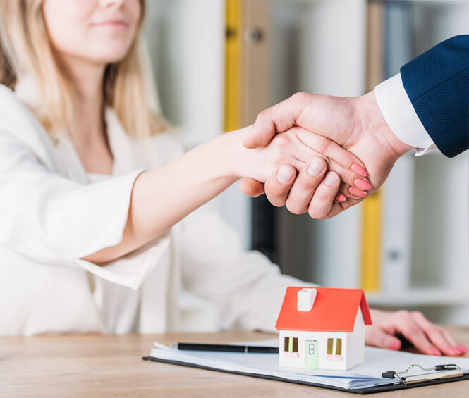 partial view of smiling woman shaking hands with realtor near house model on table