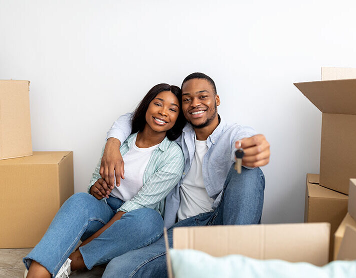 House ownership. Loving black couple holding keys, sitting among cardboard boxes and hugging after moving in new apartment. Insurance, real estate, mortgage concept